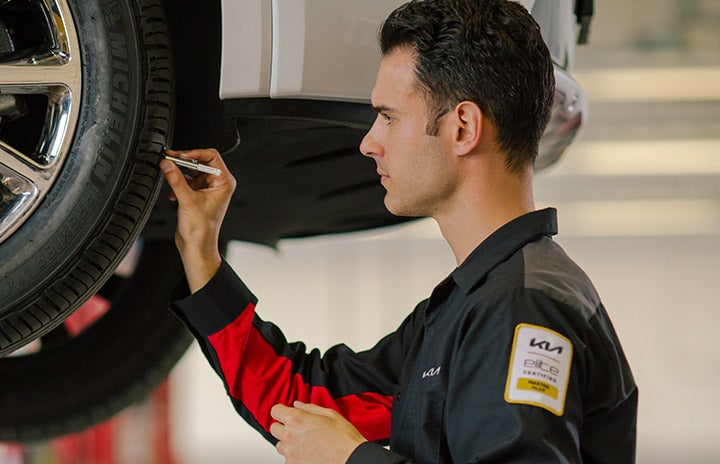 Mechanic in a black and red uniform measures tire tread of a car on a lift with a gauge.