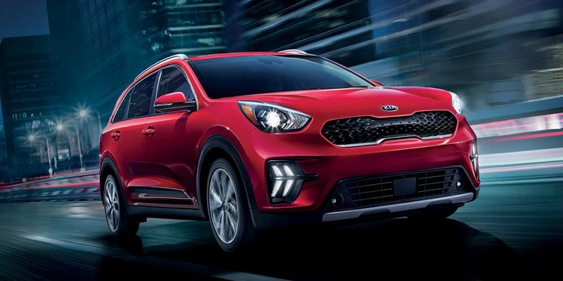 A red Kia crossover SUV with headlights on, speeding down a city street at night.