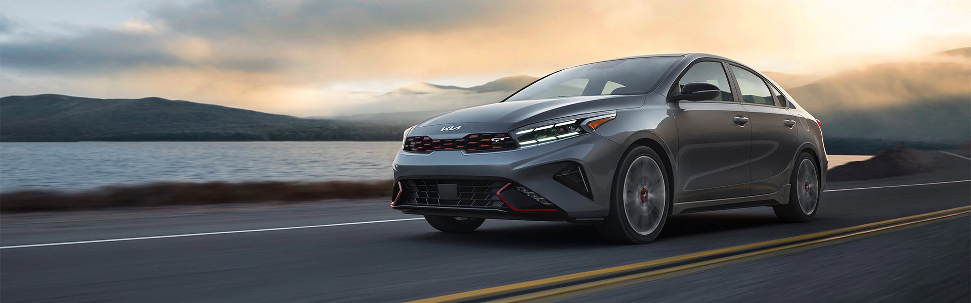 A gray Kia Forte GT driving on a road next to a lake with mountains in the background under a cloudy sky at sunset.