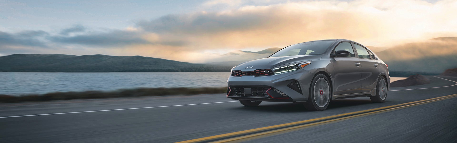 A grey Kia Forte GT sedan drives along a winding road with a lake and mountains under a cloudy sunset sky.