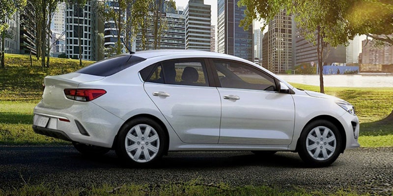 A white Kia Rio sedan parked on a gravel path with a grassy hill and city skyline in the background.