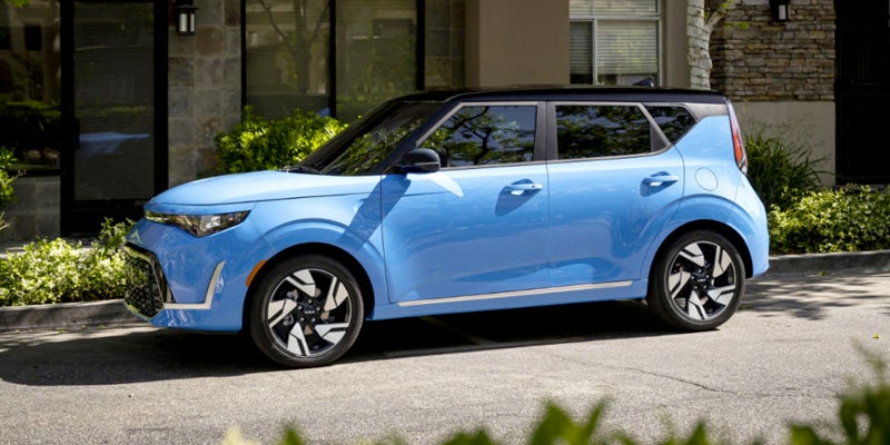 Light blue Kia Soul parked angled left, with a black roof, in front of a modern building with stone siding.
