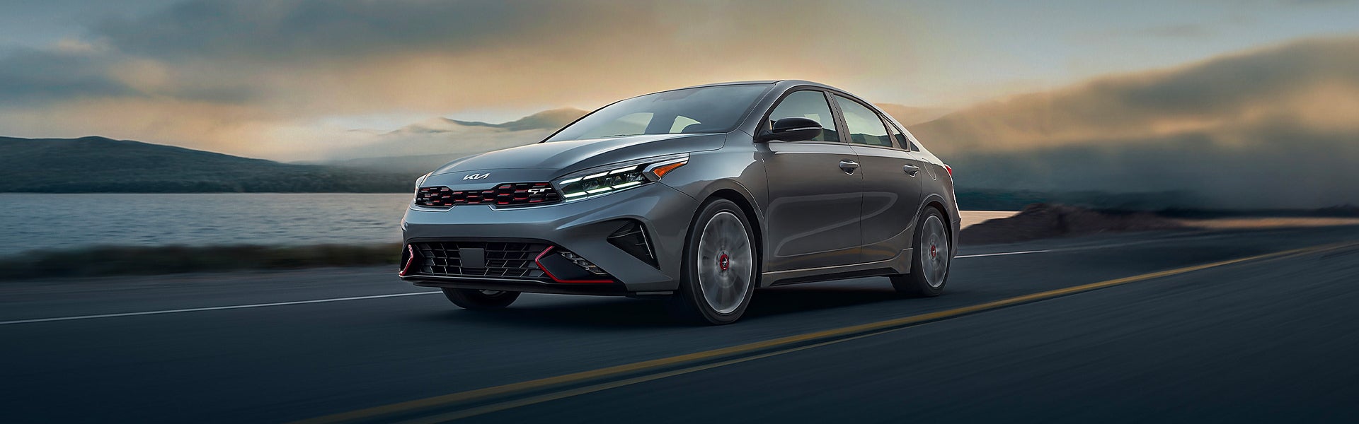 A silver Kia Forte GT on a road beside a lake with mountains in the background under a cloudy sunset.