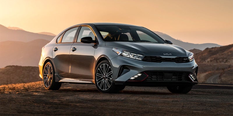 Gray Kia Forte sedan parked on a dirt road, angled slightly left, with a sunset over mountains in the background.