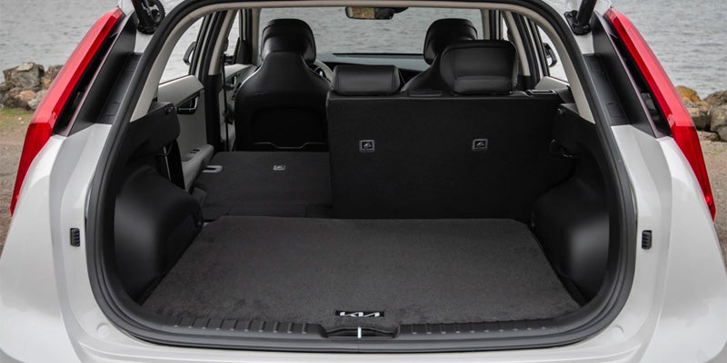 The open trunk of a light gray Kia, with half the back seats folded down, extending the dark gray carpeted floor.