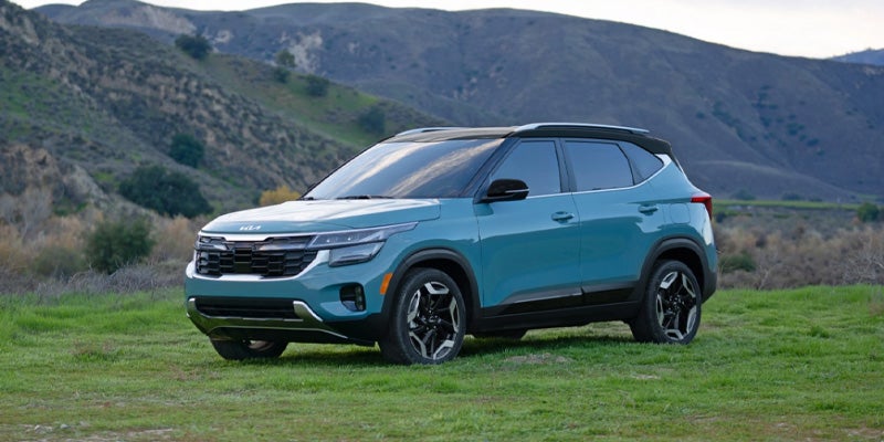A teal Kia Seltos SUV parked on light green grassy terrain with brown hills in the background under a cloudy sky.