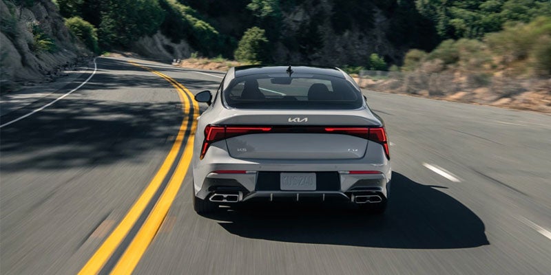 Rear view of a light gray Kia K5 sedan driving on a winding road with trees and hills in the background.