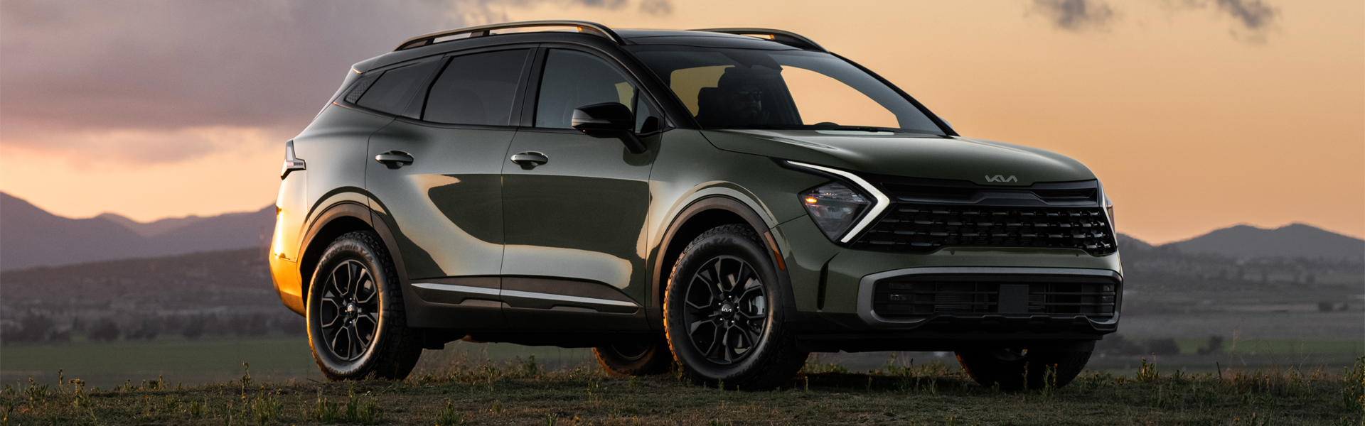A dark green Kia Sportage X-Pro is parked on a grassy hill, facing right, with golden sunset light on its side.