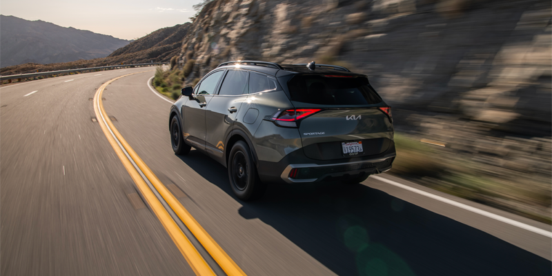 Rear view of a dark grey Kia Sportage driving quickly on a winding road with mountains in the background.