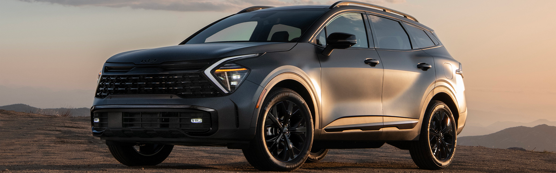A dark gray Kia Sportage SUV with black wheels glows in warm sunset light against a mountainous, hazy background.