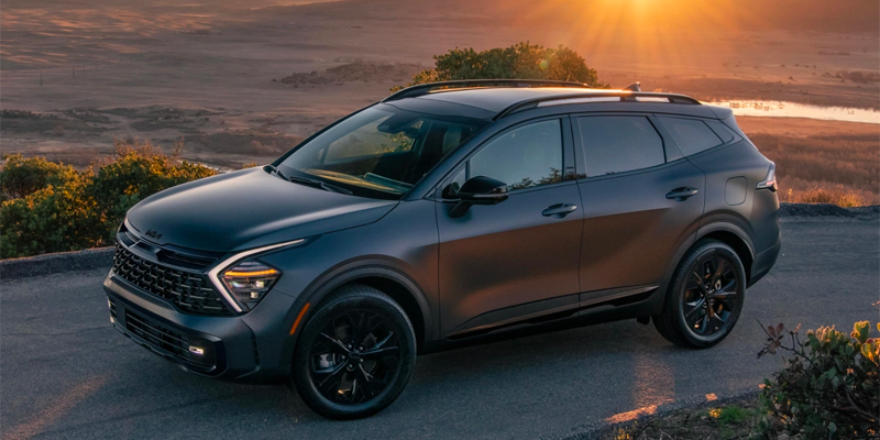 A dark matte gray Kia Sportage SUV with black wheels on a road overlooking a landscape with a bright sunset.