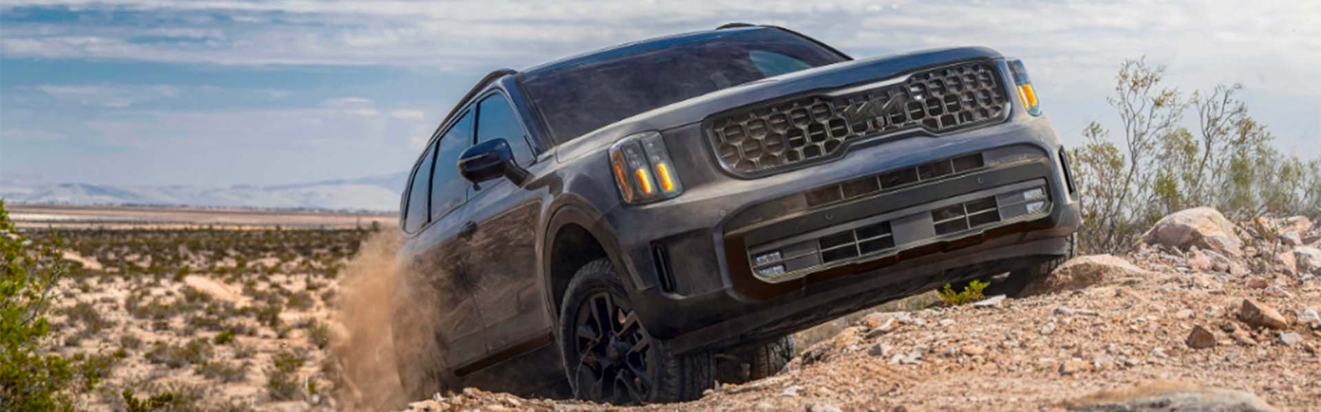 A dark gray SUV kicking up dust while driving on a rocky desert trail with mountains in the background.