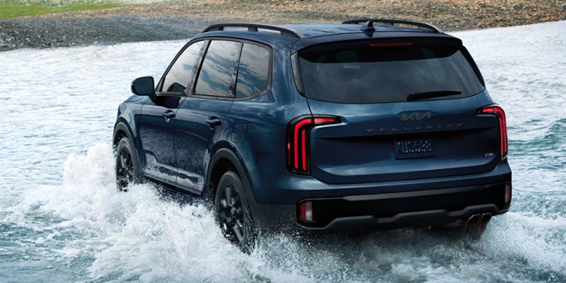 Dark blue Kia Telluride SUV driving through shallow water, creating splashes, with its rear visible.