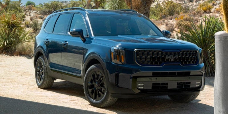 A dark blue Kia Telluride SUV parked on a dirt road in a desert landscape with scrub brush.