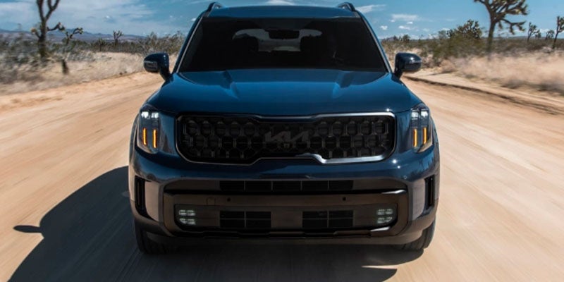 Front view of a dark blue Kia Telluride driving on a dirt road, kicked-up dust visible behind it.