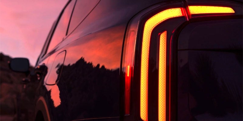Glowing red-orange taillights on a dark vehicle at dusk, reflecting a fiery sunset and distant trees.