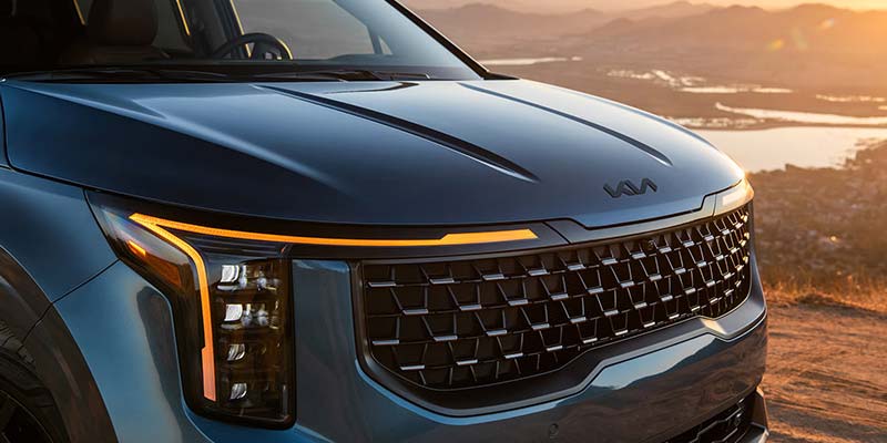 Kia Carnival SUV drives toward a sunset with a scenic mountain backdrop.