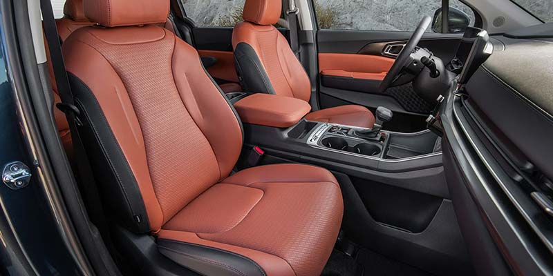 The interior of a Kia Carnival shows brown leather seats and a modern dashboard with a steering wheel.