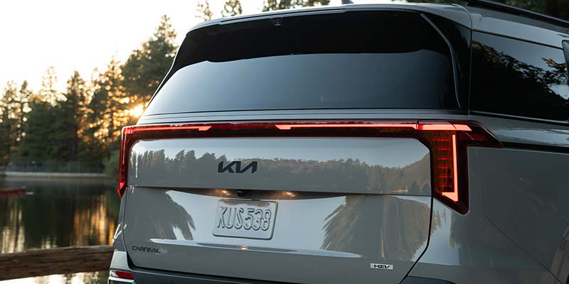 Rear view of a light Kia Carnival parked outdoors, its full-width taillight illuminated red.