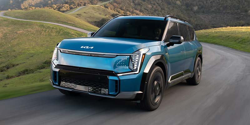 Kia EV9 drives along a winding road through a grassy, hilly landscape.