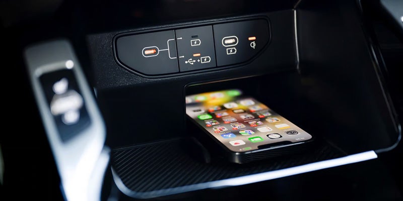 A car interior shot showing an iPhone charging wirelessly in a cubby below USB-C, USB-A and wireless charging ports.