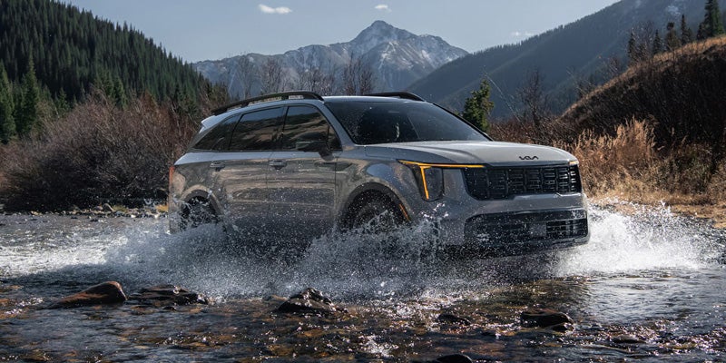 Kia Sorento SUV drives through a shallow river, splashing water, with mountains in the background.