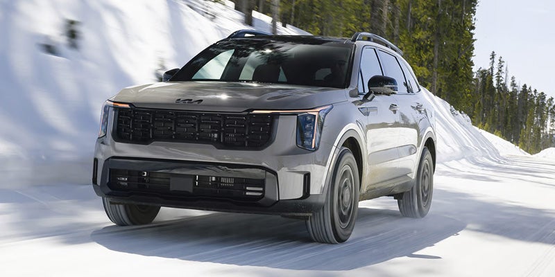 SUV with roof rails drives on a snow-covered road past snowbanks and pine trees.