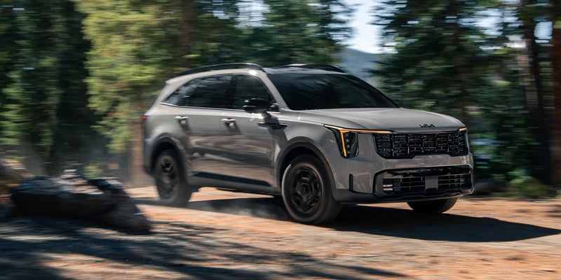 Kia Telluride SUV driving on a dirt road, kicked up dust behind it, with blurred trees in the background.