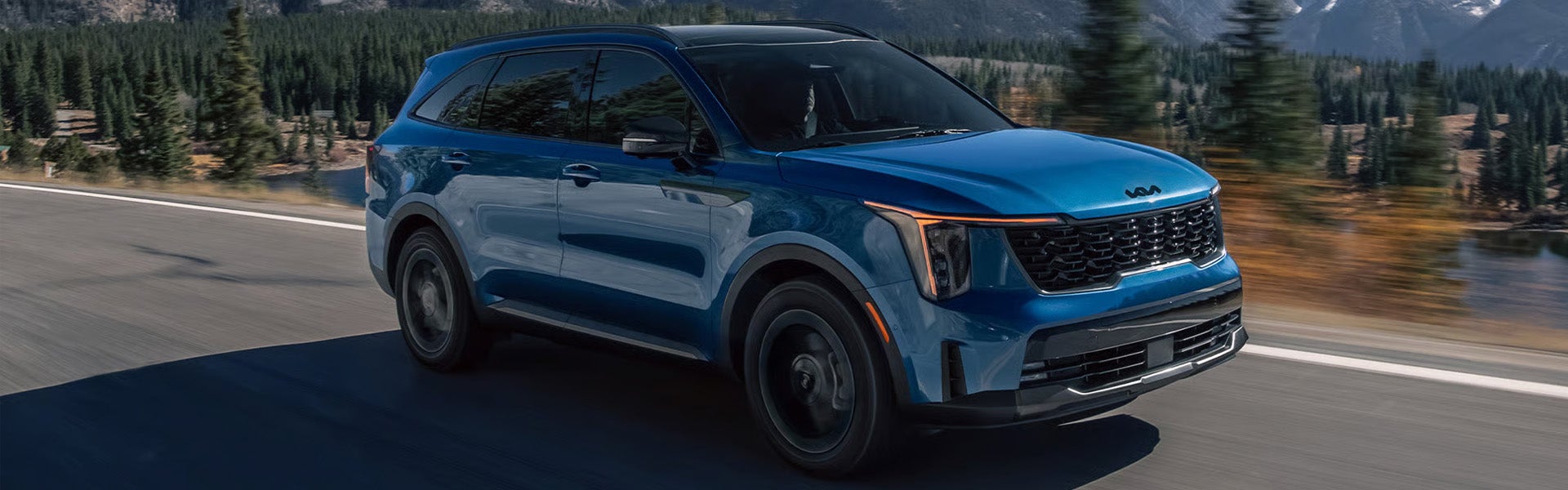 Kia Sorento drives on a country road with a mountainous, forested landscape and lake in the background.