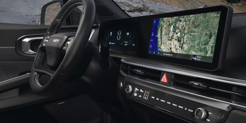 Driver's view of a modern car dashboard with a steering wheel showing the Kia logo and a large touchscreen display.