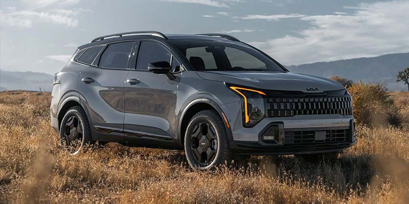 A grey Kia Sportage SUV parked in a dry grassy field with distant hills under a partly cloudy sky.