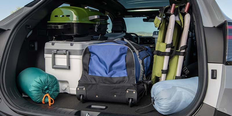 Car trunk packed with camping gear, including a green portable grill, cooler, luggage, sleeping bags, and chairs.