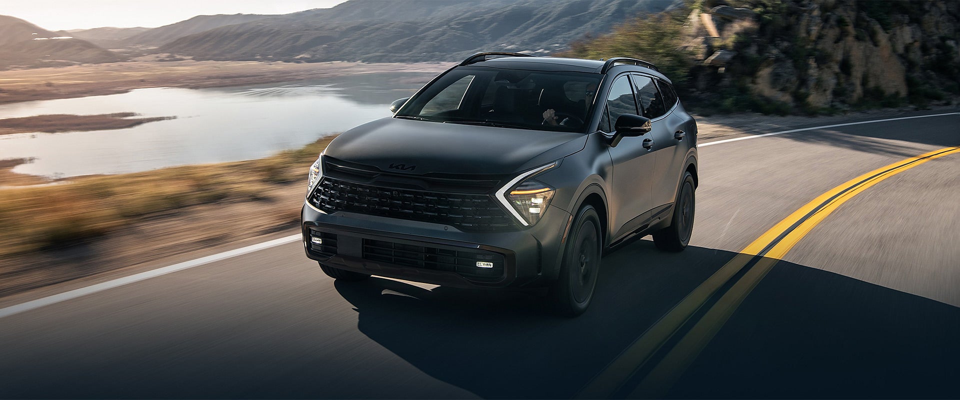 A dark gray SUV drives on a winding mountain road alongside a lake and hills, with motion blur.