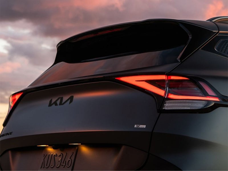 Rear view of a dark grey Kia Sportage X-Line, with bright red, boomerang-shaped LED taillights, at dusk.