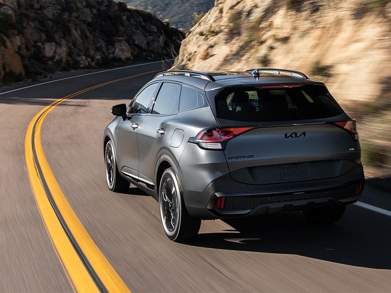 Rear view of a dark gray Kia Sportage Hybrid driving on a winding road with blurred mountain backdrop.